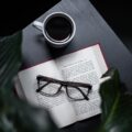 A serene setting with an open book, eyeglasses, and a steaming cup of coffee on a dark table.